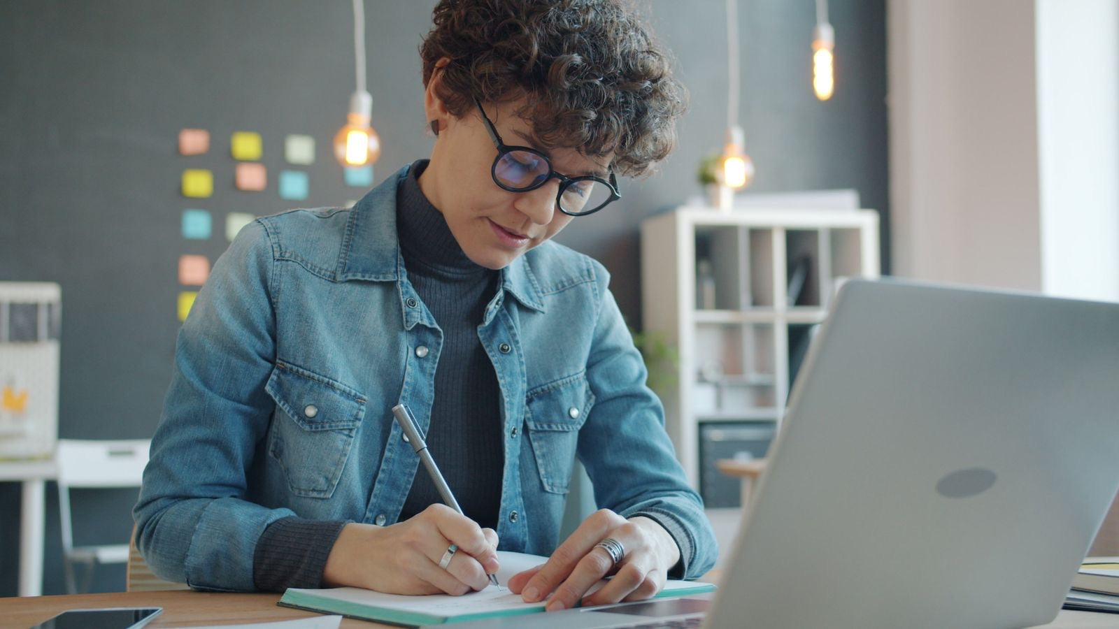 A project professional writes study notes at a laptop in an office, preparing for the new PMP exam.